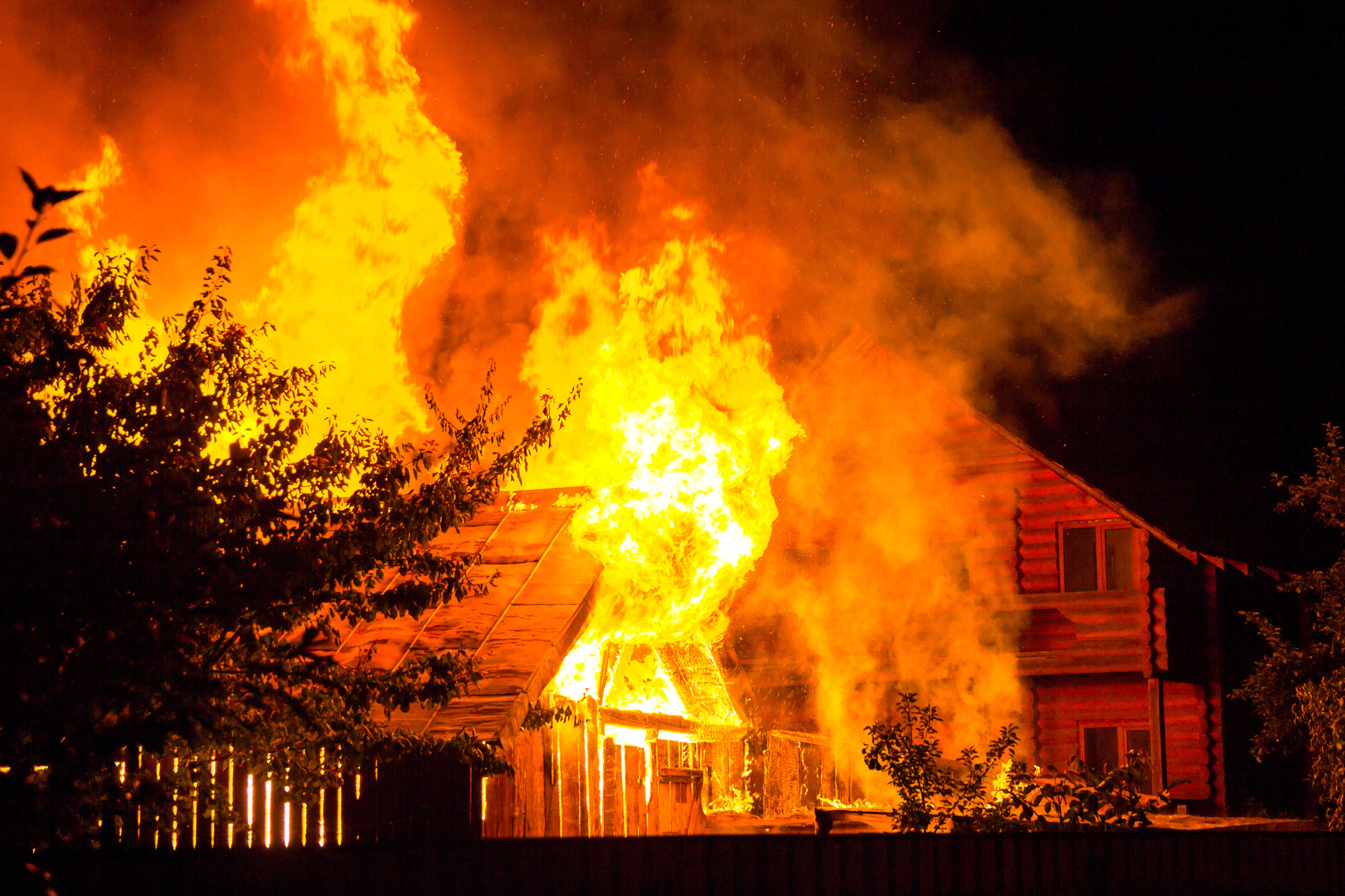 Brennendes Holzhaus bei Nacht. Helle orangefarbene Flammen und dichter Rauch steigen unter dem Ziegeldach auf, im Hintergrund zeichnen sich Baumsilhouetten und die Häuser der Nachbarn ab.