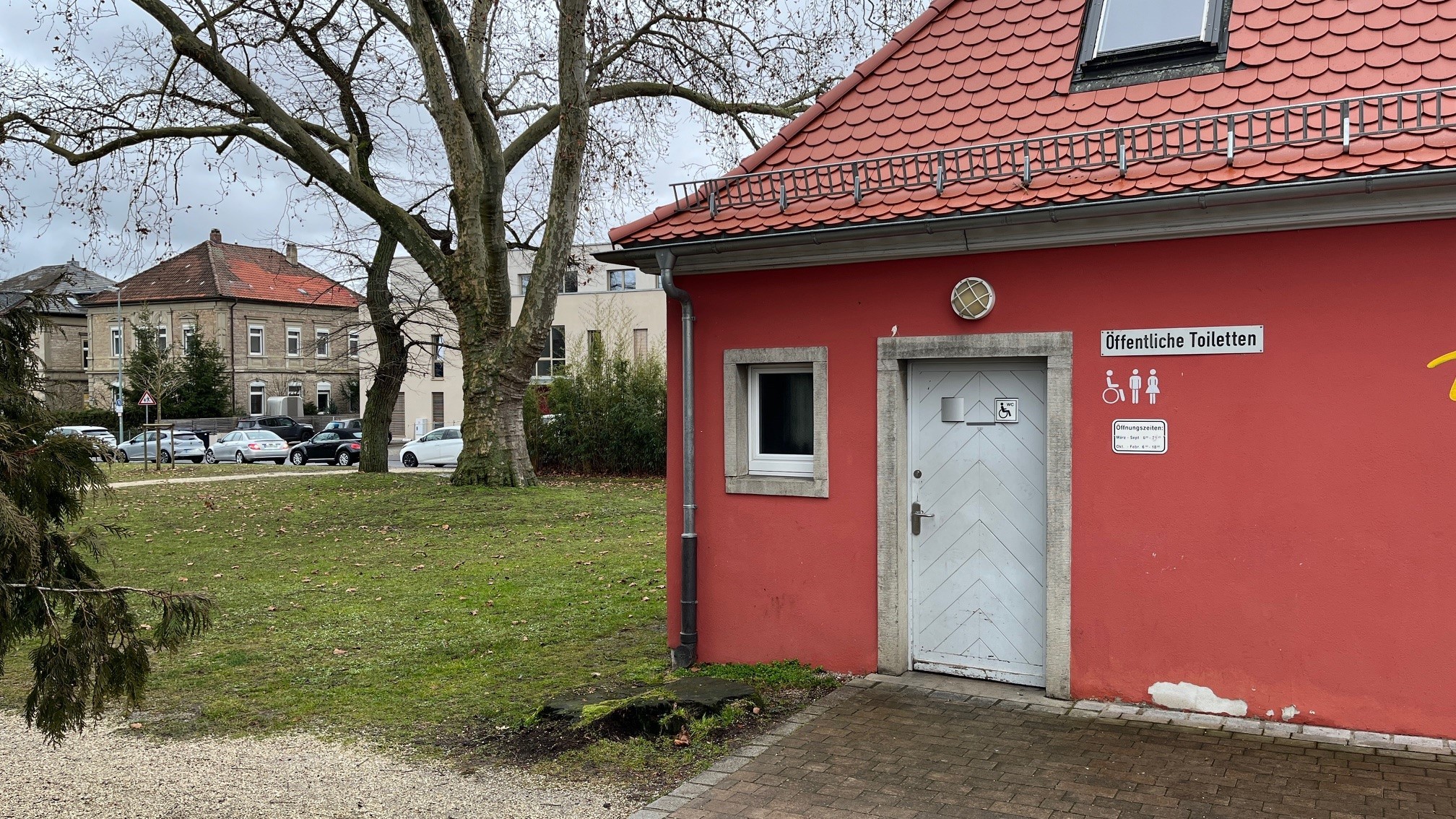 Exterior view of the public toilet in the rose garden. Signs next to the white door indicate the toilet.