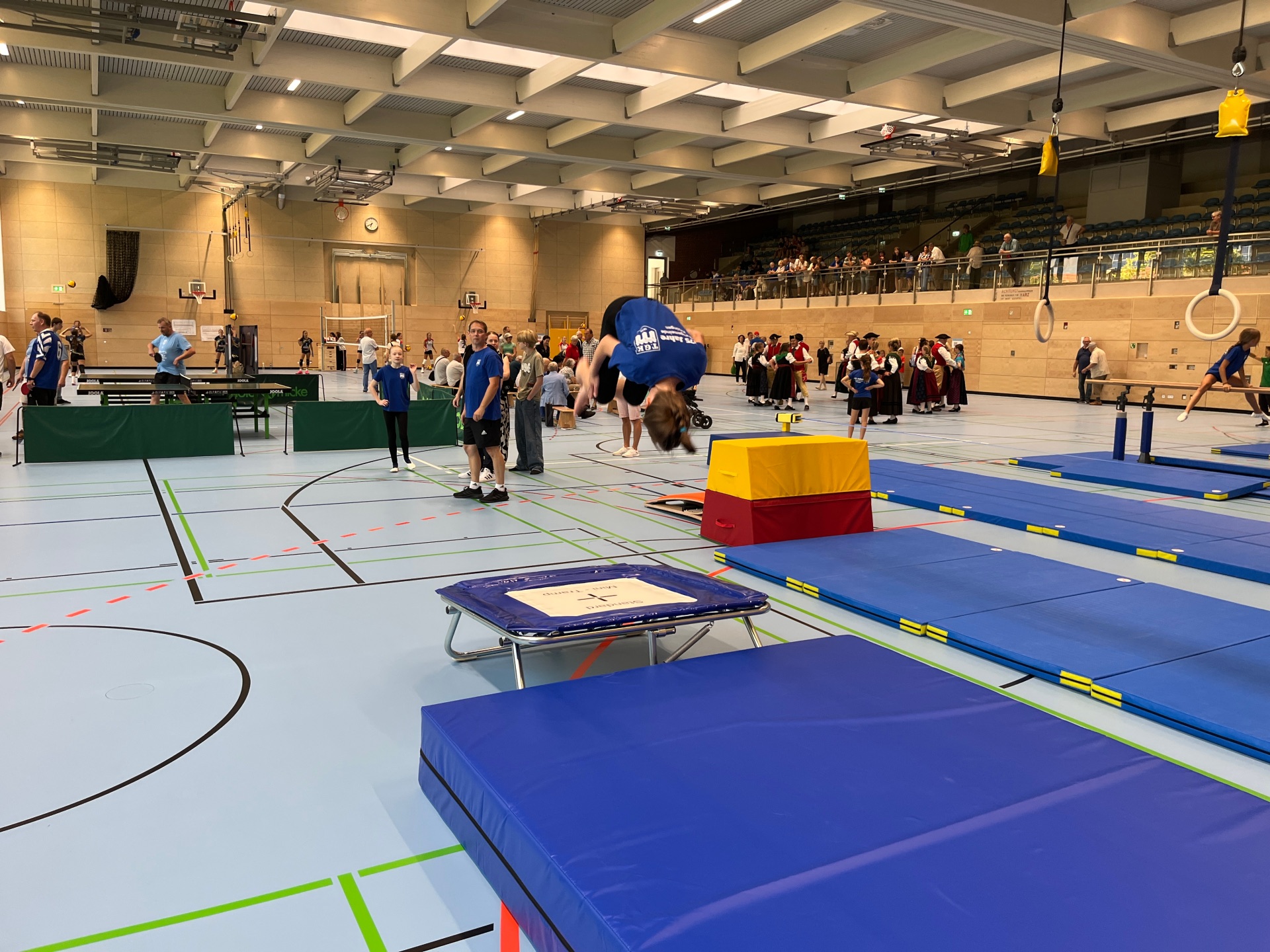 Ein Mädchen macht einen Salto nach einem Sprung vom Trampolin auf eine Matte. Im Hintergrund spielen Leute Tischtennis und die Volkstanzgruppe führt einen Tanz auf.